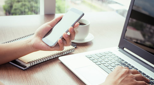 hand holding a phone and typing on a laptop